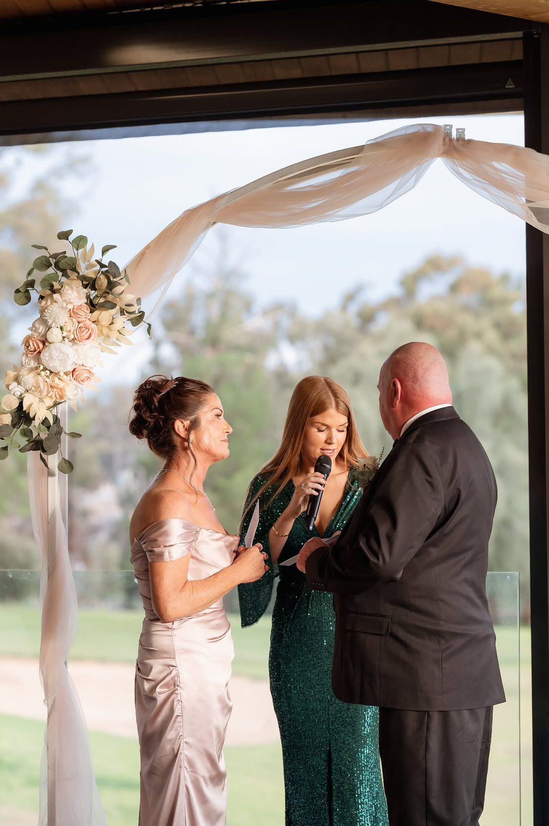 Katie in a green sequinned dress reading into a microphone between the couple under a draped floral arbour at Shepparton Golf Club, with the fairways behind