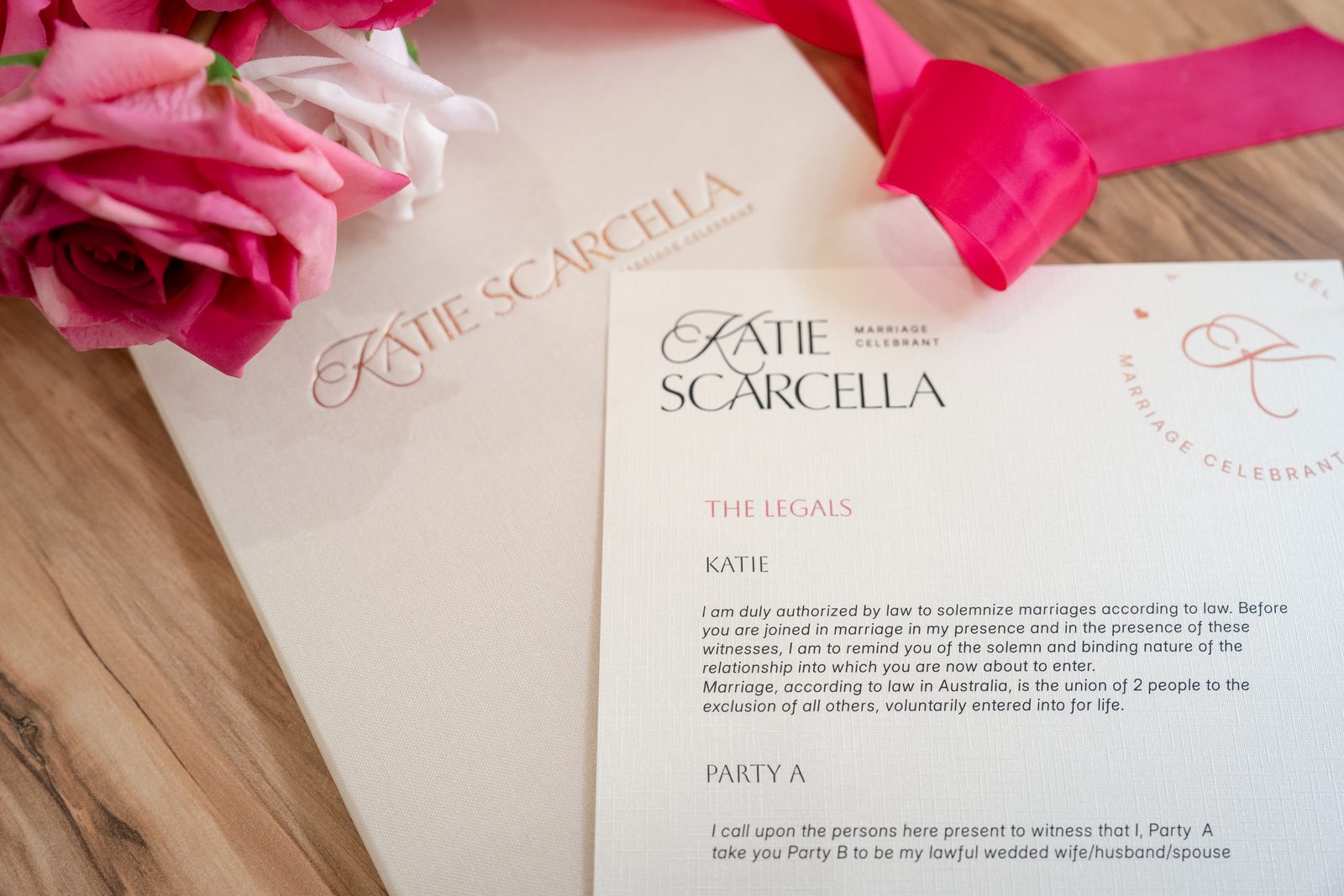Katie Scarcella’s ‘The Legals’ card showing the legal words for the celebrant and the couple, on a cream folder beside pink roses and a hot pink ribbon