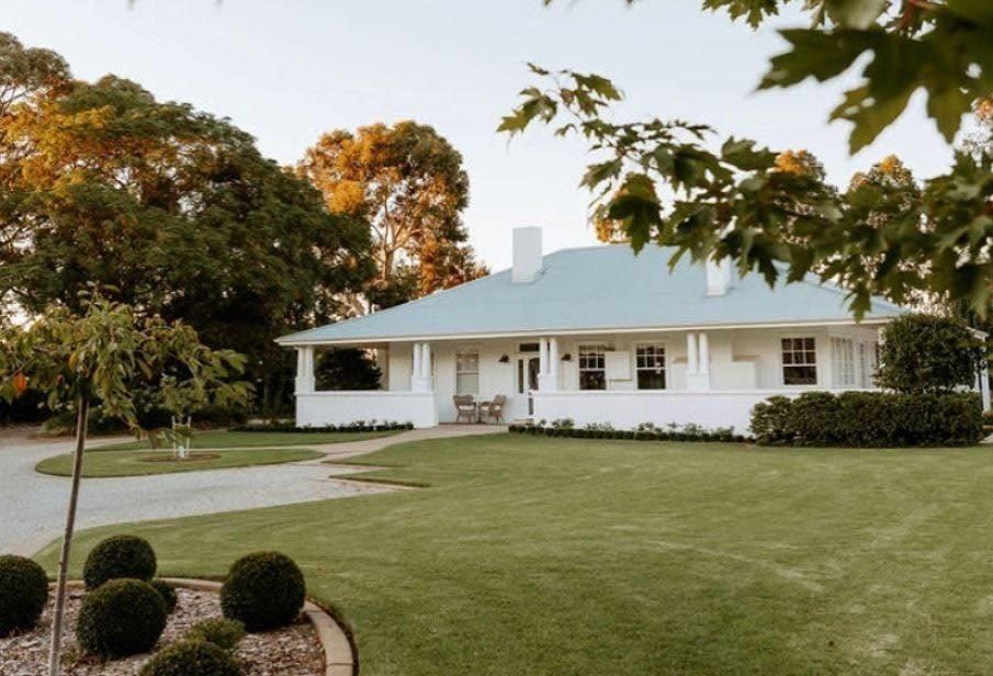 The restored white 1930s farmhouse at Modern Country Estate, with its wide verandah, manicured lawn and curved driveway