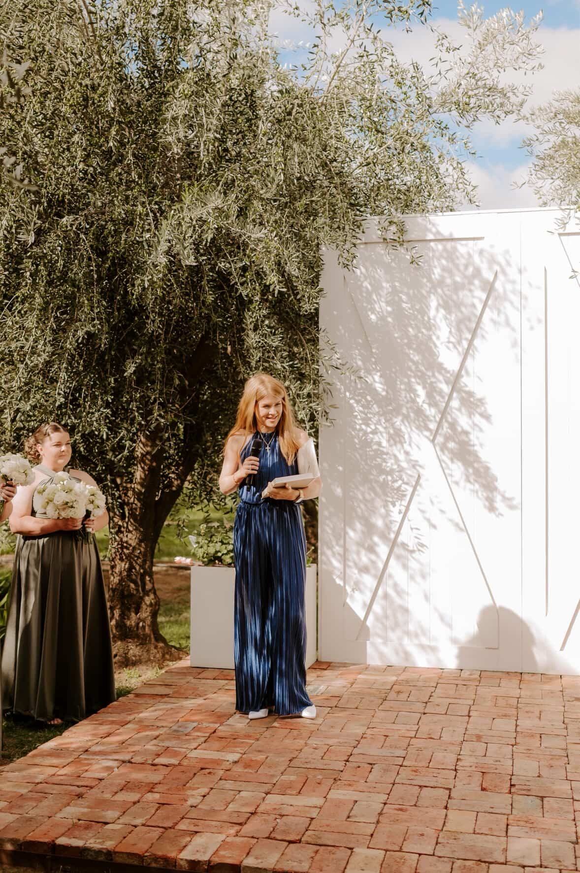 Katie smiling with a microphone and ceremony book in front of the white barn doors and olive tree at Olivehouse, with a bridesmaid holding white flowers