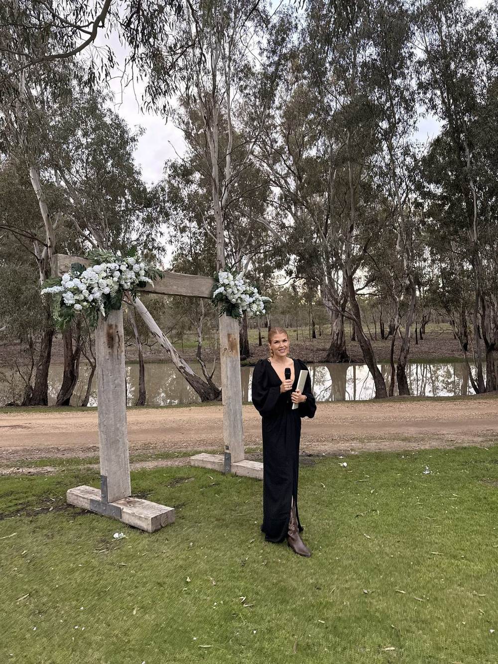 Katie smiling with a microphone and ceremony book beside a timber arbour with white flowers, in front of the lagoon and river red gums at The Paddock at Ulupna