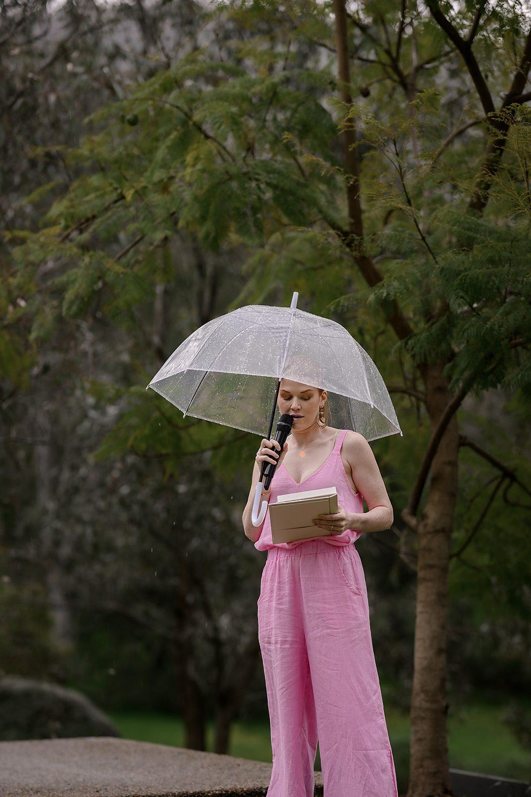 Katie in a pink jumpsuit holding a clear umbrella and microphone, reading a ceremony in the rain at The Trawool Estate