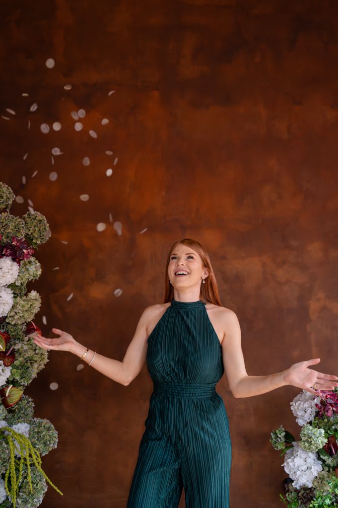 Katie laughing as confetti falls, in a green jumpsuit against a rust-coloured wall with flowers