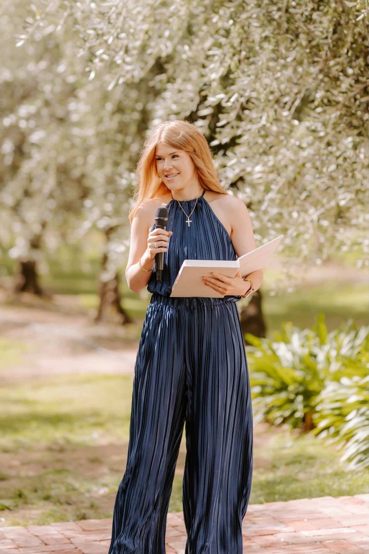 Katie smiling with a microphone and ceremony folder in an olive grove