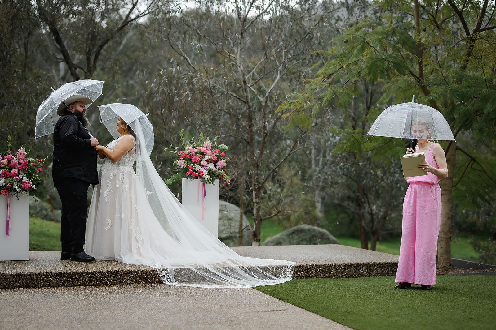 Katie in a pink jumpsuit leading a ceremony under clear umbrellas as the couple hold hands in the rain