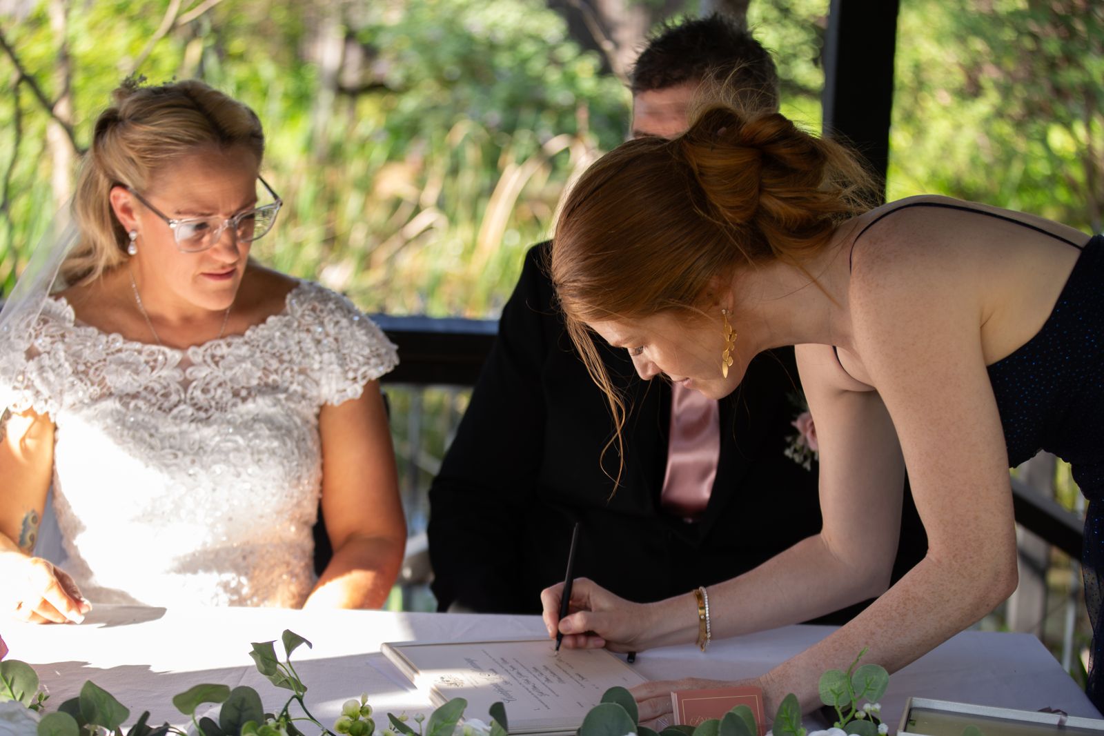 Katie signing the marriage certificate beside the bride at an outdoor ceremony